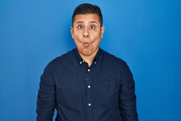 Hispanic young man standing over blue background making fish face with lips, crazy and comical gesture. funny expression.