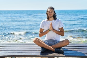 Young hispanic man doing yoga exercise sitting on bench at seaside