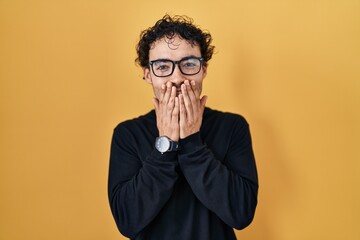 Hispanic man standing over yellow background laughing and embarrassed giggle covering mouth with hands, gossip and scandal concept
