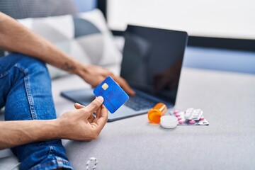 Young hispanic man using laptop and credit card sitting on sofa at home