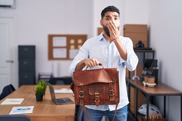 Hispanic man with beard working at the office holding briefcase covering mouth with hand, shocked...