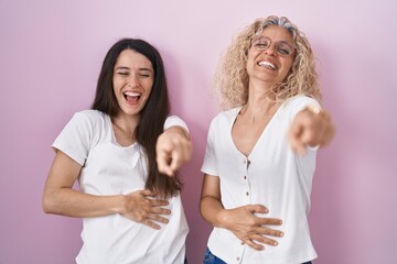 Mother and daughter standing together over pink background laughing at you, pointing finger to the camera with hand over body, shame expression