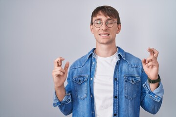 Caucasian blond man standing wearing glasses gesturing finger crossed smiling with hope and eyes closed. luck and superstitious concept.