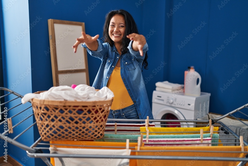 Poster Young asian woman hanging clothes at clothesline looking at the camera smiling with open arms for hug. cheerful expression embracing happiness.