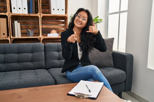 Young asian woman at consultation office smiling doing talking on the telephone gesture and pointing to you. call me.