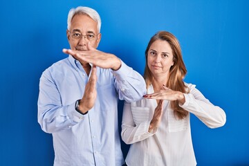 Middle age hispanic couple standing over blue background doing time out gesture with hands,...
