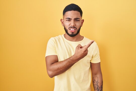 Young hispanic man standing over yellow background pointing aside worried and nervous with forefinger, concerned and surprised expression