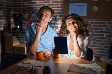 Middle age hispanic couple using touchpad sitting on the table at night with hand on chin thinking about question, pensive expression. smiling with thoughtful face. doubt concept.