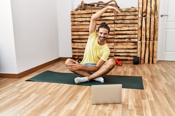 Handsome hispanic man stretching looking at tutorial on laptop at the gym