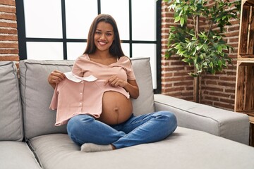 Young latin woman pregnant holding baby clothes at home