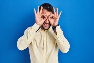 Handsome hispanic man standing over blue background doing ok gesture like binoculars sticking tongue out, eyes looking through fingers. crazy expression.