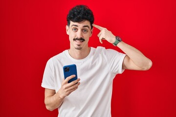 Hispanic man using smartphone over red background smiling pointing to head with one finger, great idea or thought, good memory