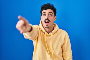 Hispanic man standing over blue background pointing with finger surprised ahead, open mouth amazed expression, something on the front