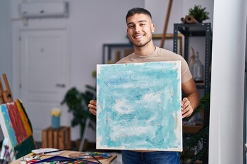 Young hispanic painter man holding canvas smiling with a happy and cool smile on face. showing...