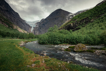 Naklejka na ściany i meble Mountain river with ice cold water of Briksdalsbreen glacier in the mountains of Jostedalsbreen national park in Norway, moody atmosphere, rocks in the water