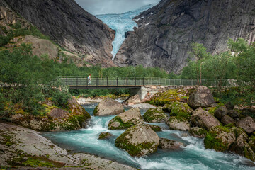 Mountain river with ice cold water of Briksdalsbreen glacier in the mountains of Jostedalsbreen national park in Norway, woman hiking over bridge - 559152136