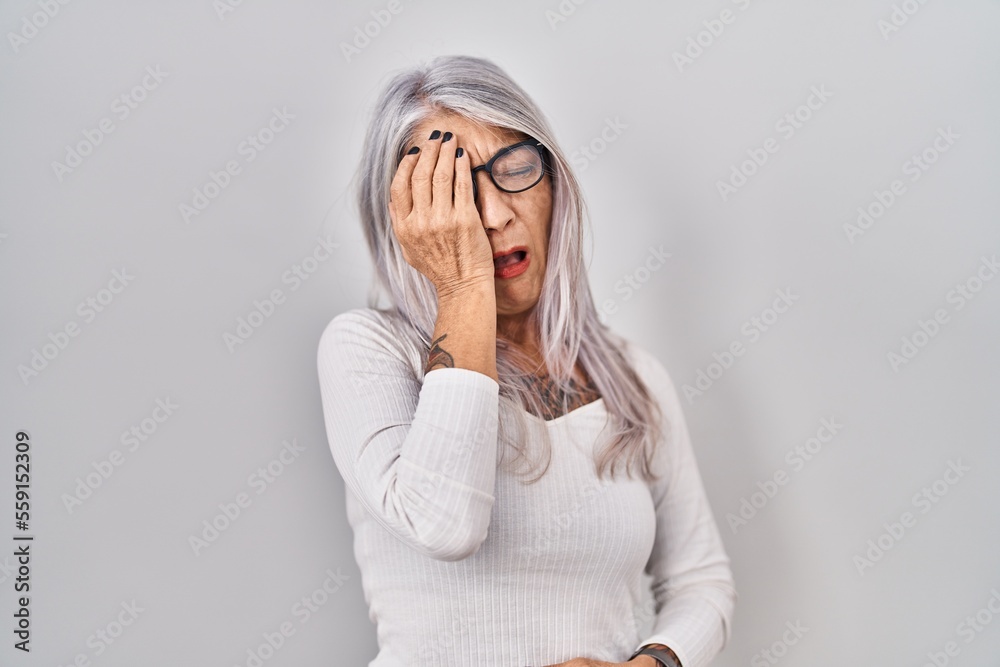 Wall mural Middle age woman with grey hair standing over white background yawning tired covering half face, eye and mouth with hand. face hurts in pain.