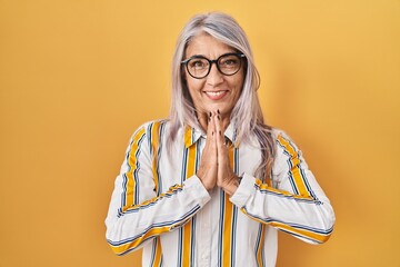 Middle age woman with grey hair standing over yellow background wearing glasses praying with hands together asking for forgiveness smiling confident.
