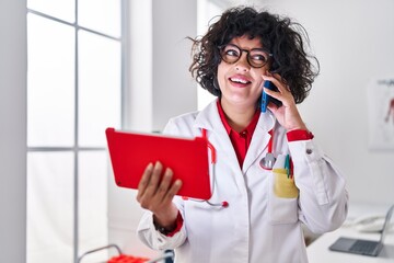 Young beautiful hispanic woman doctor talking on smartphone using touchpad at clinic
