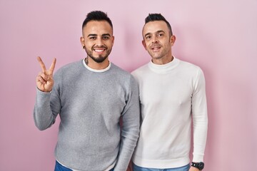 Homosexual couple standing over pink background showing and pointing up with fingers number two while smiling confident and happy.