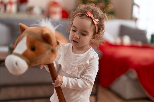 Adorable caucasian girl playing with horse toy standing by christmas tree at home