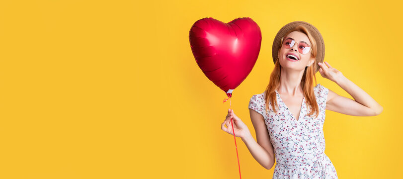 cheerful girl in straw hat and sunglasses hold love heart balloon on yellow background. Woman isolated face portrait, banner with copy space.