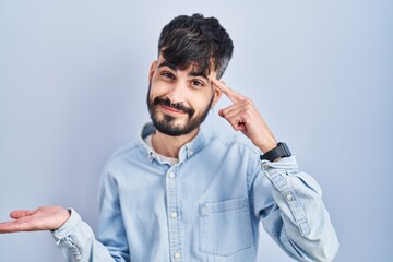 Fototapeta premium Young hispanic man with beard standing over blue background confused and annoyed with open palm showing copy space and pointing finger to forehead. think about it.