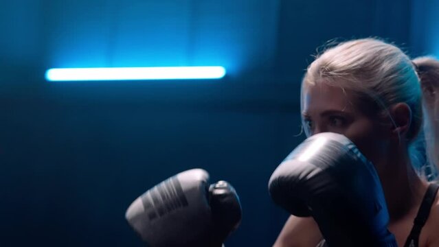 Female fighter training in dark gym with blue light. Woman in black boxing gloves practices punches on punching bag held by a trainer. Kickboxing workout. Close up. Slow motion ready, 4K at 59.94fps.
