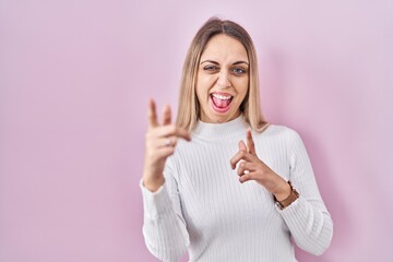 Young blonde woman wearing white sweater over pink background pointing fingers to camera with happy and funny face. good energy and vibes.