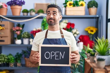 Hispanic man with beard working at florist holding open sign smiling looking to the side and staring away thinking.