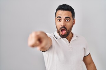 Young hispanic man with beard wearing casual clothes over white background pointing with finger surprised ahead, open mouth amazed expression, something on the front