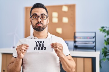 Young hispanic man with beard holding you are fired banner at the office puffing cheeks with funny...