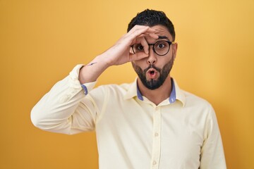 Hispanic young man wearing business clothes and glasses doing ok gesture shocked with surprised face, eye looking through fingers. unbelieving expression.