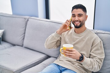 Fototapeta premium Young arab man drinking coffee and talking on the smartphone at home