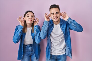 Fototapeta na wymiar Young hispanic couple standing over pink background trying to hear both hands on ear gesture, curious for gossip. hearing problem, deaf