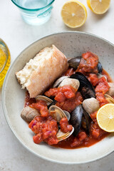 Beige plate with steamed mussels and littleneck clams in thick tomato broth with ciabatta and lemon, vertical shot, middle close-up