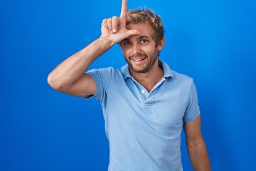 Caucasian man standing over blue background making fun of people with fingers on forehead doing...