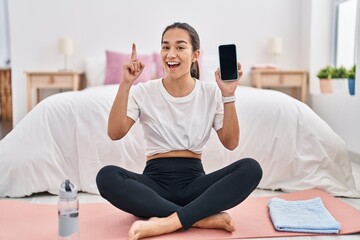 Fototapeta na wymiar Young south asian woman doing yoga mat holding smartphone showing screen smiling with an idea or question pointing finger with happy face, number one