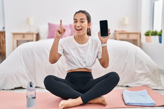 Young south asian woman doing yoga mat holding smartphone showing screen smiling with an idea or question pointing finger with happy face, number one