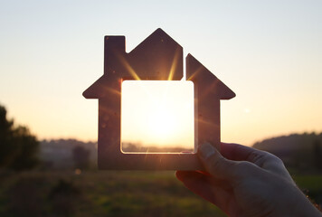 A hand holding a wooden house against the sky and the sun in nature. Idea of solar energy, and...