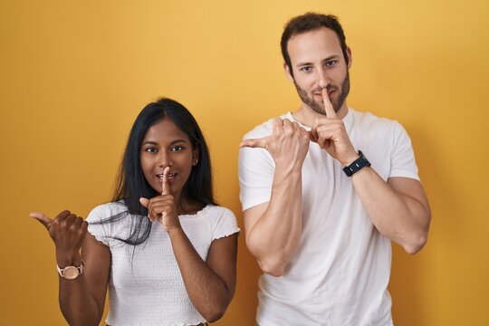 Interracial couple standing over yellow background asking to be quiet with finger on lips pointing with hand to the side. silence and secret concept.