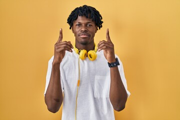 Young african man with dreadlocks standing over yellow background pointing up looking sad and...