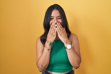 Brunette woman standing over yellow background laughing and embarrassed giggle covering mouth with...