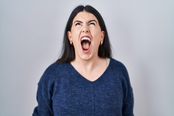 Young brunette woman standing over isolated background angry and mad screaming frustrated and furious, shouting with anger. rage and aggressive concept.