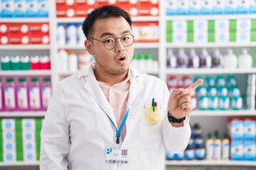 Chinese young man working at pharmacy drugstore surprised pointing with finger to the side, open mouth amazed expression.