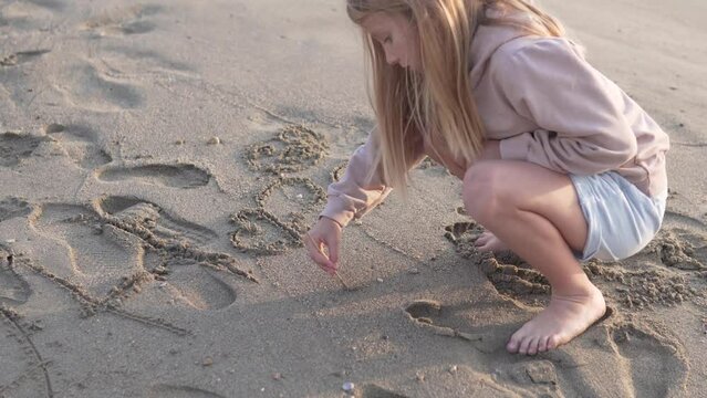  Little girl draws with a stick on a sandy beach at sunset.