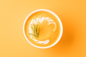 Thick butternut squash bisque in white bowl garnished with heavy cream and rosemary over orange background. Bowl of pumpkin cream soup. View overhead. Selective focus