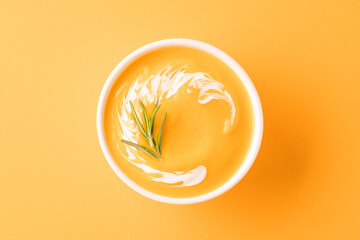 Thick butternut squash bisque in white bowl garnished with heavy cream and rosemary over orange...