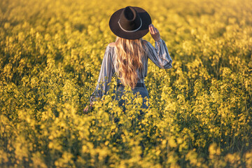 Blonde woman standing in the blooming yellow rapeseed field at sunset. Copy space.Color of vintage and retro tones.