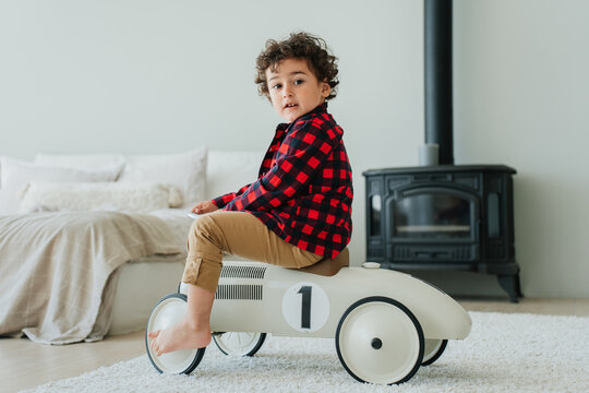 Curly caucasian little boy in plaid shirt and casual pants sitting on toy car at bedroom looks at camera. Handsome child received a present from parents. Cute toddler playing at home. Childhood. № one